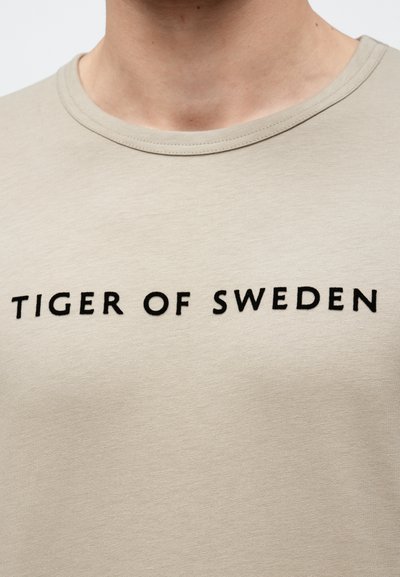 Svetlo béžové tričko s okrúhlym výstrihom. Na hrudi sa nachádza nápis "TIGER OF SWEDEN" v tučných čiernych písmenách. Hladká textúra.