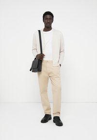 Beige cardigan med knapper, hvit t-skjorte, beige bukser, svarte sko og en svart skulderveske, mot en enkel hvit bakgrunn.