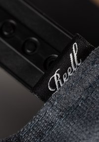 Graues Stoffetikett mit weißem "Reell"-Text, angebracht an einem strukturierten Kleidungsstück, neben einem schwarzen Material mit kreisförmigem Detail und Verschluss.