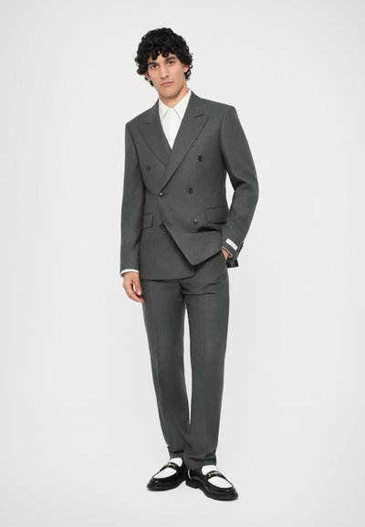 Abito doppiopetto in tessuto grigio scuro, caratterizzato da rever appuntiti, due tasche laterali, abbinato a una camicia bianca e scarpe nere.