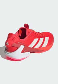 Červená sportovní bota s síťovaným svrškem, třemi bílými pruhy a výraznou bílou podrážkou. Obsahuje unikátní design paty a poutko.