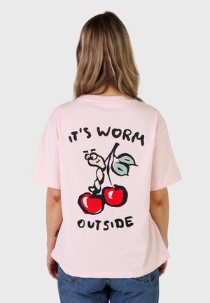 Kvinne med langt hår iført en rosa T-skjorte som viser en mark på to kirsebær med blader og teksten "It's worm outside" på baksiden.