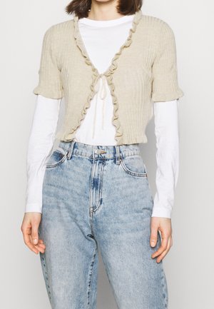 Cardigan beige côtelé court avec des bords volants, noué à l'avant, manches courtes, porté par-dessus une chemise blanche à manches longues et associé à un jean bleu clair.