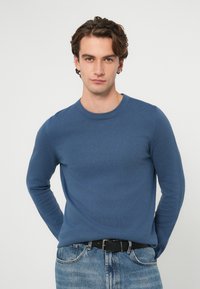 Niebieski sweter z dzianiny z okrągłym dekoltem, dopasowany krój, długie rękawy, zestawiony z wyblakłymi niebieskimi dżinsami i czarnym paskiem.