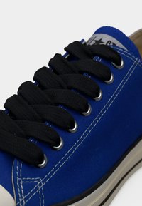 Μπλε πάνινο αθλητικό παπούτσι με χοντρές μαύρες κορδόνια περασμένες μέσα από ασημί ιμάντες και λευκά διακοσμητικά ράμματα στην πλευρά.