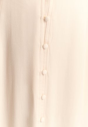 Heledat beeži pluus, millel on sile tekstuur, vertikaalsed õmblused ja ees ümmarguste kangaga kaetud nööpidega nööpide plaat.