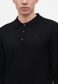 Homme portant un polo noir à manches longues avec trois boutons et un logo brodé discret sur la poitrine.