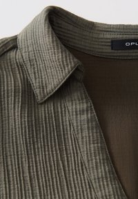 Chemise à col en tissu côtelé texturé vert olive avec doublure intérieure marron foncé, montrant des coutures détaillées et une partie de l'étiquette au niveau du col.