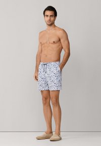 Oberkörperfreier Mann in weißen Badeshorts mit blauem Palmenmuster und beigen Slip-on-Schuhen, stehend vor einem schlichten hellgrauen Hintergrund.