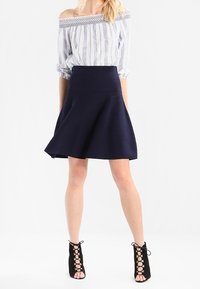 Blouse rayée bleue claire et blanche à épaules dénudées, associée à une jupe évasée bleu marine. Des bottines à lacets noires complètent la tenue.