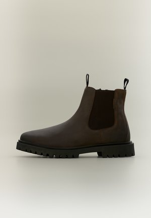 Dunkelbrauner Leder-Chelsea-Boot mit schwarzem elastischem Seitenteil, schwarzen Zuglaschen und dicker schwarzer Profilsohle vor neutralem Hintergrund.