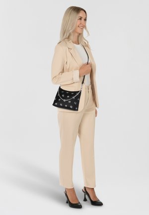 Completo beige su misura con giacca aderente, abbinato a scarpe con tacco nere. Una borsa nera con tracolla a catena presenta un motivo di piccole icone.