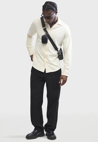 Chemise à col crème avec une texture verticale, pantalon en velours côtelé noir et sac bandoulière noir. Des chaussures noires arrondies complètent la tenue.
