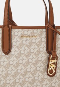 Vaaleanruskea ja beige kuvioitu Michael Kors -laukkua, jossa on ruskeat nahkahandat ja kultainen logo-riipus kiinnitettynä yhteen handoista.