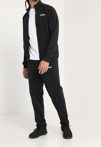Chándal negro hecho de poliéster suave, con una chaqueta con cremallera y pantalones con cintura elástica. Logo blanco de Adidas en la chaqueta y en los pantalones.