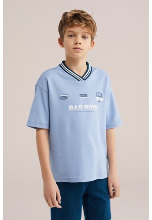 Niño con cabello castaño claro, vestido con una camiseta estilo fútbol azul claro con el texto "Bad Boys" y pantalones azul marino, posando frente a un fondo neutro.