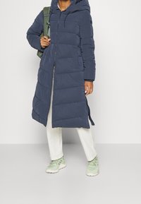 Mørkeblå puffervinterjakke med hætte, der har lynlås-lukning, vandret syninger og to sidelommer. Bæres med off-white bukser og grønne sneakers.