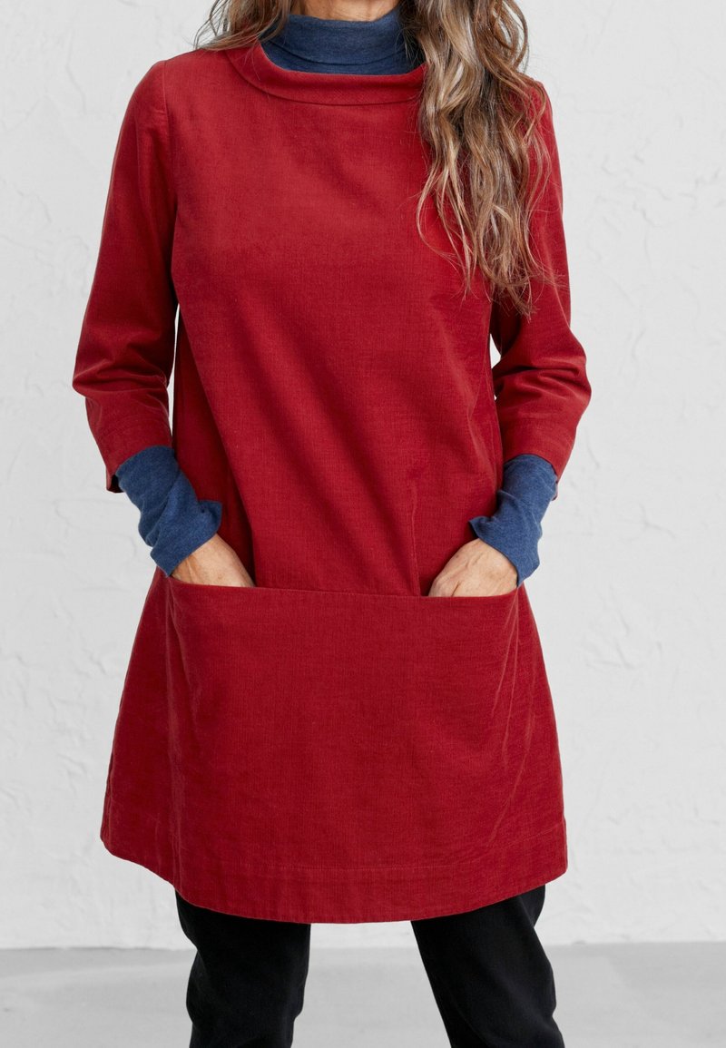 Robe en velours côtelé rouge avec des manches longues, un décolleté large et de grandes poches avant. Portée par-dessus un col roulé bleu, mettant en valeur des textures superposées.