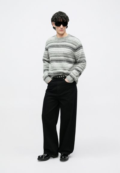 Sivi i crni prugasti džemper, široke crne hlače, crni pojas s okruglim detaljima i sjajne crne cipele. Glatka tekstura kroz cijelu odjeću.