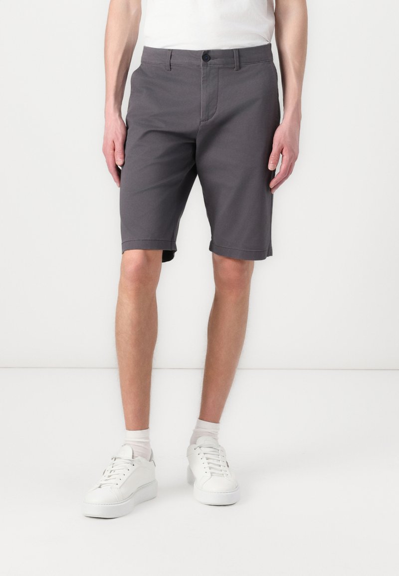 Graue Baumwollshorts mit taillierter Passform, ausgestattet mit einem Knopfverschluss, Seitentaschen und aufgerolltem Saum, getragen mit weißen Sneakers.