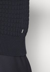 Gros plan d'un pull noir en maille torsadée avec bord-côte à l'ourlet et petite étiquette en tissu "BOSS" sur le côté, sur fond clair.