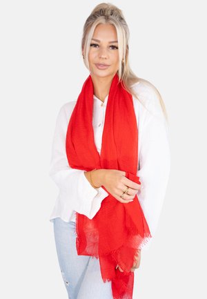 Red textured scarf draped over hands, featuring frayed edges and lightweight fabric. Worn over a white shirt and light blue jeans.