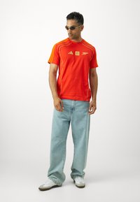 Rode sportieve t-shirt met gele strepen en logo's, gecombineerd met lichtblauwe wijde broek en witte sneakers. Man draagt een zonnebril.