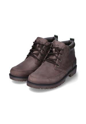Botas de cuero marrón hasta el tobillo con cordones en la parte frontal, puntera redonda y suela texturizada. Presentan un collar acolchado y una cinta para facilitar el calzado.