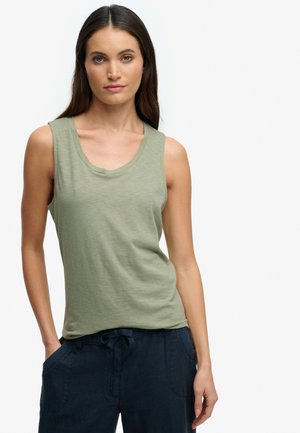 Femme aux longs cheveux bruns portant un haut sans manches vert clair et un pantalon bleu foncé, debout avec une main dans la poche sur fond blanc.