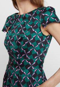 Hihaton mekko tummanvihreästä kankaasta, jossa on geometrinen kuvio, mustilla ja vaaleanpunaisilla yksityiskohdilla sekä muotoonommeltu yläosa, jossa on laskosyksityiskohta.