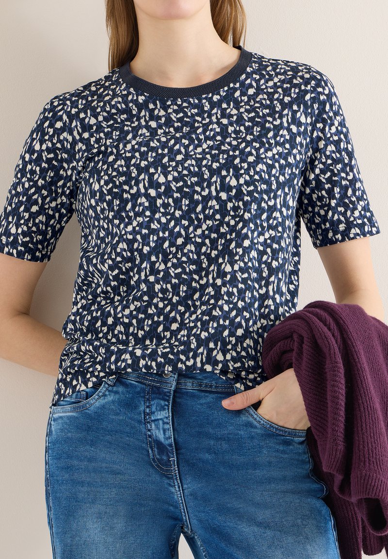 Femme portant un haut à manches courtes bleu marine et blanc à motifs avec un jean bleu, tenant un pull en tricot violet foncé plié dans une main.