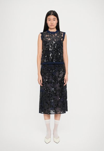 Vestido azul marino con tela transparente superpuesta con lentejuelas. El diseño incluye un escote alto y sisas con flecos. Detalle de cintura delgada visible.