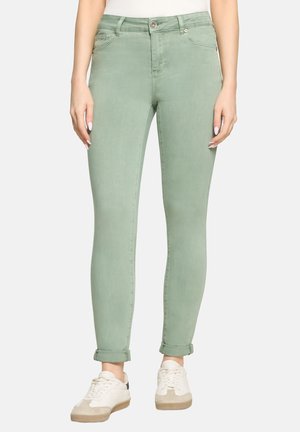 Jean slim vert clair avec ourlets retroussés, porté avec des baskets blanches sur une personne debout contre un fond uni.