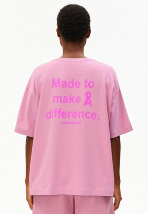 Persoon met een loszittend roze shirt met de tekst "Made to make a difference." en kleiner "ARMEDANGELS" gedrukt op de achterkant.
