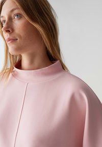 Roze oversized top met een hoge kraag, voorzien van een gladde textuur en subtiel nadenontwerp, ontworpen voor een ontspannen pasvorm.