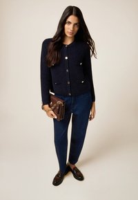 Cardigan in tessuto navy con bottoni dorati, due tasche anteriori, abbinato a pantaloni blu scuro con delicati luccichii e borsa marrone.