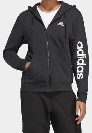 Personne portant un sweat à capuche zippé noir Adidas avec un logo blanc sur la poitrine et la manche, associé à un pantalon noir.
