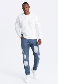 Sudadera blanca con patrón texturizado, combinada con jeans azules desgastados que presentan grandes desgastes, y zapatillas blancas. Look simple y casual.