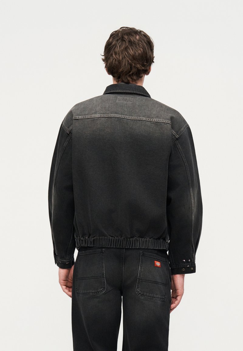 Uomo con capelli castani che indossa una giacca di denim nera lavata e jeans abbinati, visto da dietro su uno sfondo semplice.