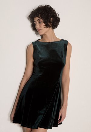 Femme aux cheveux courts et bouclés portant une robe en velours vert foncé sans manches, debout contre un mur clair uni, regardant vers le bas.
