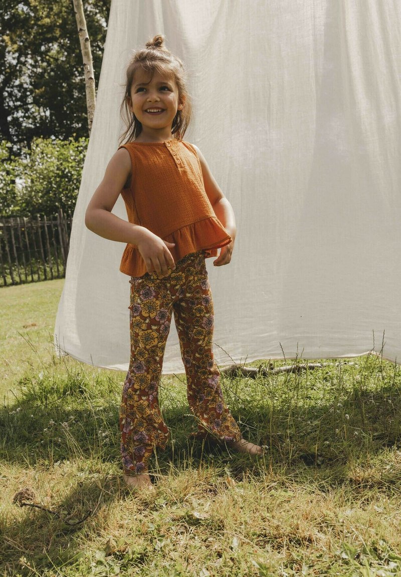 Een oranje mouwloze top met een gerimpelde onderrand en knopen aan de voorkant, gecombineerd met kleurrijke bloemenbedrukte flared broeken. Buiten op gras.