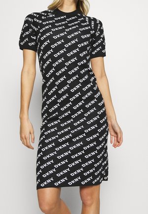 Czarna sukienka z krótkim rękawem z białymi logo DKNY drukowanymi ukośnie na tkaninie, noszona przez osobę stojącą na tle jednolitego tła.