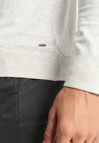 Hellgrauer Pullover mit geripptem Saum und Bündchen, versehen mit einem kleinen metallischen Logo-Tag nahe dem Saum. Der Stoff scheint weich und strukturiert zu sein.