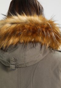 Cheveux bruns visibles au-dessus de la capuche d'une parka vert olive avec une épaisse bordure en fausse fourrure brun doré autour de la capuche.