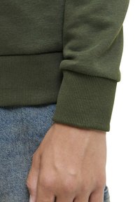 Olijfgroen sweatshirt manchet gemaakt van ribgebreid materiaal, gecombineerd met lichtblauwe denim, die textuur en een strak design toont.