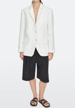 Person iført en hvid blazer med to knapper, sorte løse knælange shorts og mørkebrune sandaler med åben tå mod en ensfarvet baggrund.