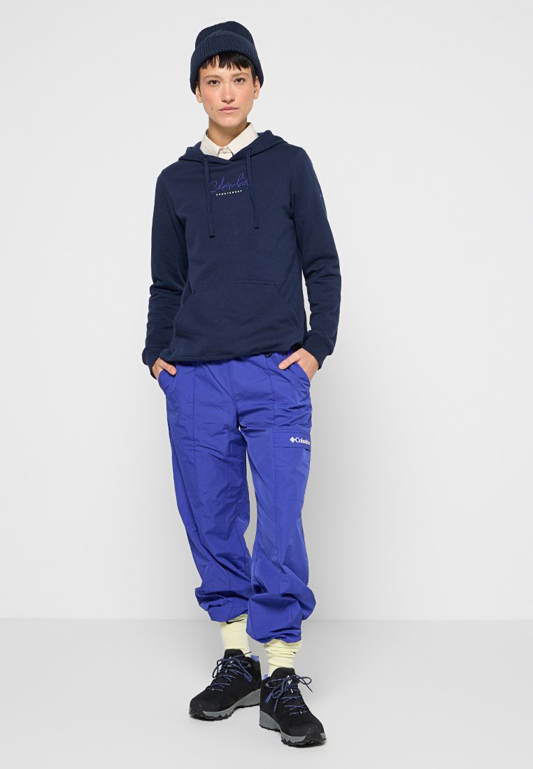 Felpa navy con tasca frontale e coulisse, abbinata a pantaloni azzurri brillanti con logo. Scarpe nere con suole testurizzate.