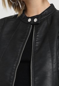 Veste en cuir noir avec un col haut, quincaillerie en argent visible et fermeture éclair à l'avant. Présente des coutures sur mesure et une texture lisse.