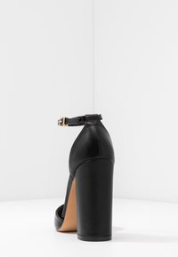 Scarpa nera con tacco alto in pelle texturizzata, con cinturino alla caviglia e tacco solido, che mostra un design minimalista.