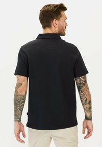 Kortærmet polo skjorte i mørkeblå med en struktureret stof, diskret krave og sideskår. Tatovering synlig på højre arm.